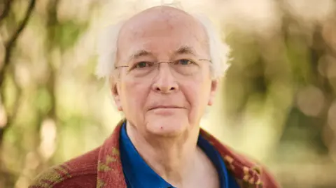 Sam Allard Philip Pullman with glasses in a brown & beige cardigan and blue shirt against trees and leaves background