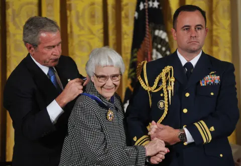 Getty Images President George W. Bush in dark suit and blue tie, putting the Presidential Medal of Freedom around Harper Lee's neck. Harper smiling, wearing glasses, dressed in black and white dog tooth outfit, holding hand of man in uniform