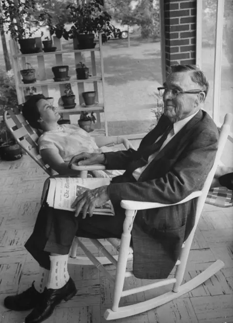 Getty Images Harper Lee sitting outside on a reclining chair in short sleeved dress looking upwards, next to her father in dark suit and glasses, with a newspaper on his lap. Plants on shelf in the background